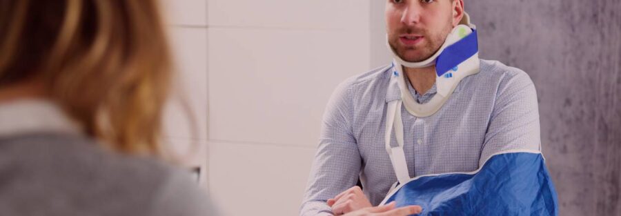 Mężczyzna w kołnierzu ortopedycznym i temblaku, siedzący przy szklanym stole, rozmawia z kobietą w biurze. W tle widać białą szafkę oraz dekoracyjne rośliny. Na zdjęciu znajduje się logo "Votum Odszkodowania SA".