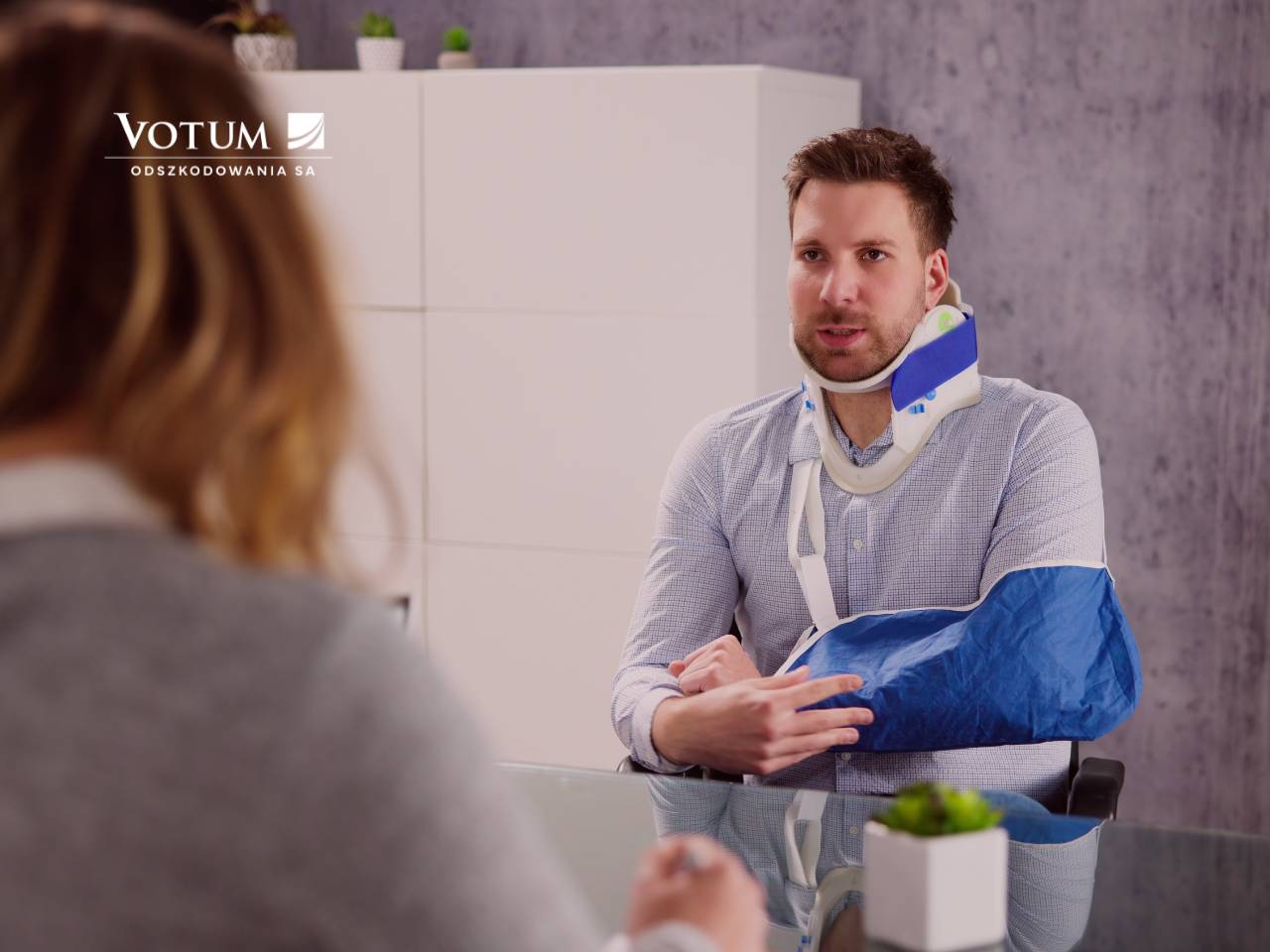 Mężczyzna w kołnierzu ortopedycznym i temblaku, siedzący przy szklanym stole, rozmawia z kobietą w biurze. W tle widać białą szafkę oraz dekoracyjne rośliny. Na zdjęciu znajduje się logo "Votum Odszkodowania SA".