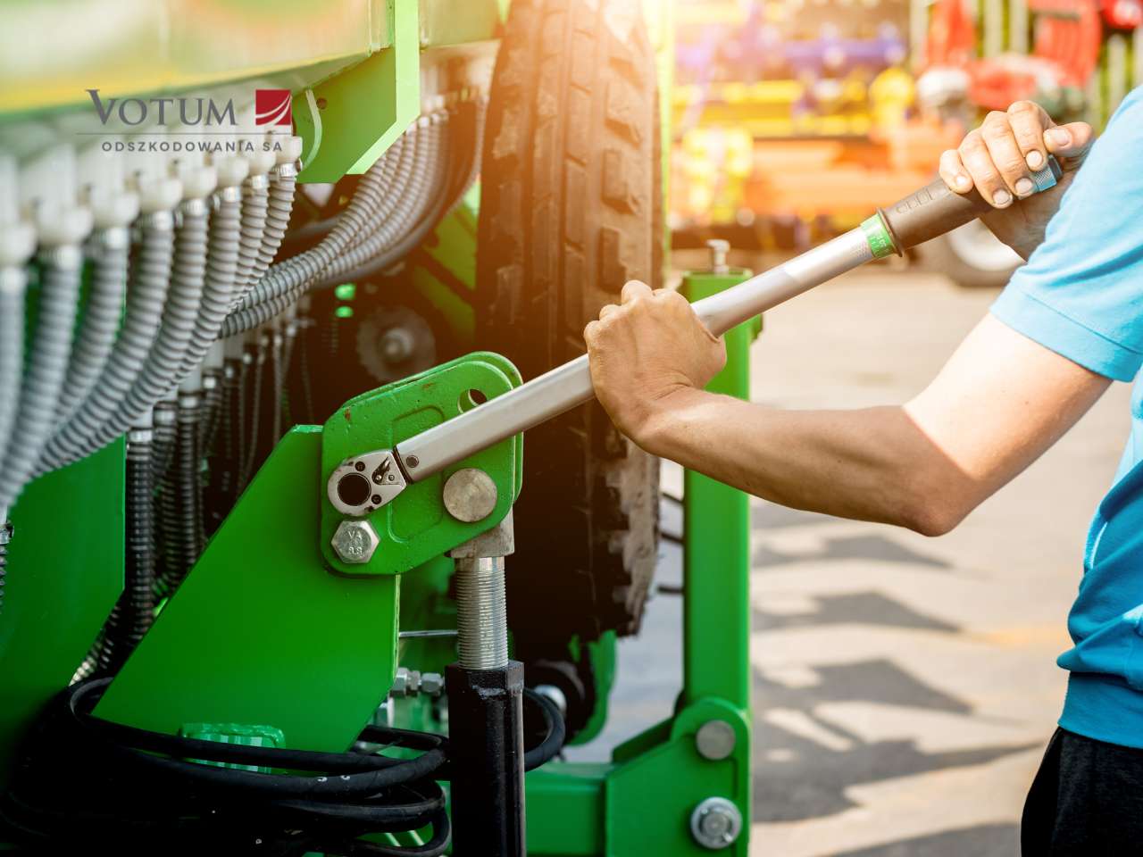 Mężczyzna w niebieskiej koszulce naprawia maszynę rolniczą, używając dużego klucza, w lewym górnym rogu widoczne logo Votum Odszkodowania SA