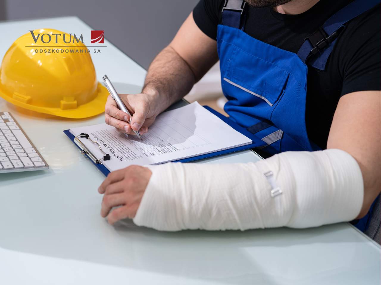 Mężczyzna w gipsie na ręku wyplnia wniosek o odszkodowanie za wypadek w pracy. obok leży żółty kask.