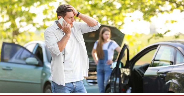 Mężczyzna rozmawia przez telefon z zmartwioną miną, stojąc obok miejsca wypadku drogowego. W tle kobieta stoi przy samochodzie z otwartą maską. W tle widać zieloną, naturalną scenerię.