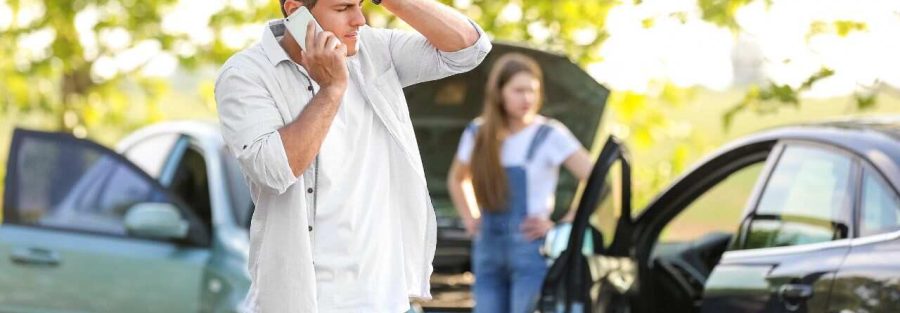 Mężczyzna rozmawia przez telefon z zmartwioną miną, stojąc obok miejsca wypadku drogowego. W tle kobieta stoi przy samochodzie z otwartą maską. W tle widać zieloną, naturalną scenerię.