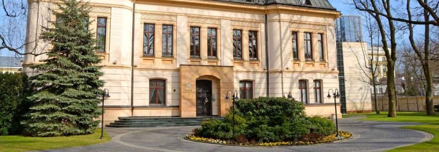 Fasada budynku Trybunału Konstytucyjnego w Warszawie otoczona drzewami, z widocznym paskiem informacyjnym „Artykuł na biznes.pap – Służebność przesyłu po wyroku TK” oraz logo Votum Odszkodowania SA.