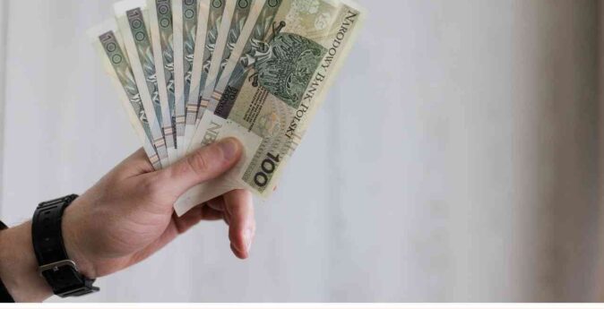 Męska dłoń trzyma wachlarz banknotów stuzłotowych na tle grafiki informacyjnej o rencie z OC sprawcy.